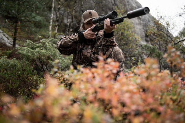 Kvinne i kamuflasjeklær sikter med en Browning X-Bolt rifle i norsk natur. X-Bolt er kjent for sin presisjon, sikkerhet og brukervennlighet, og har vært en av Norges mest populære jaktrifler i flere år.